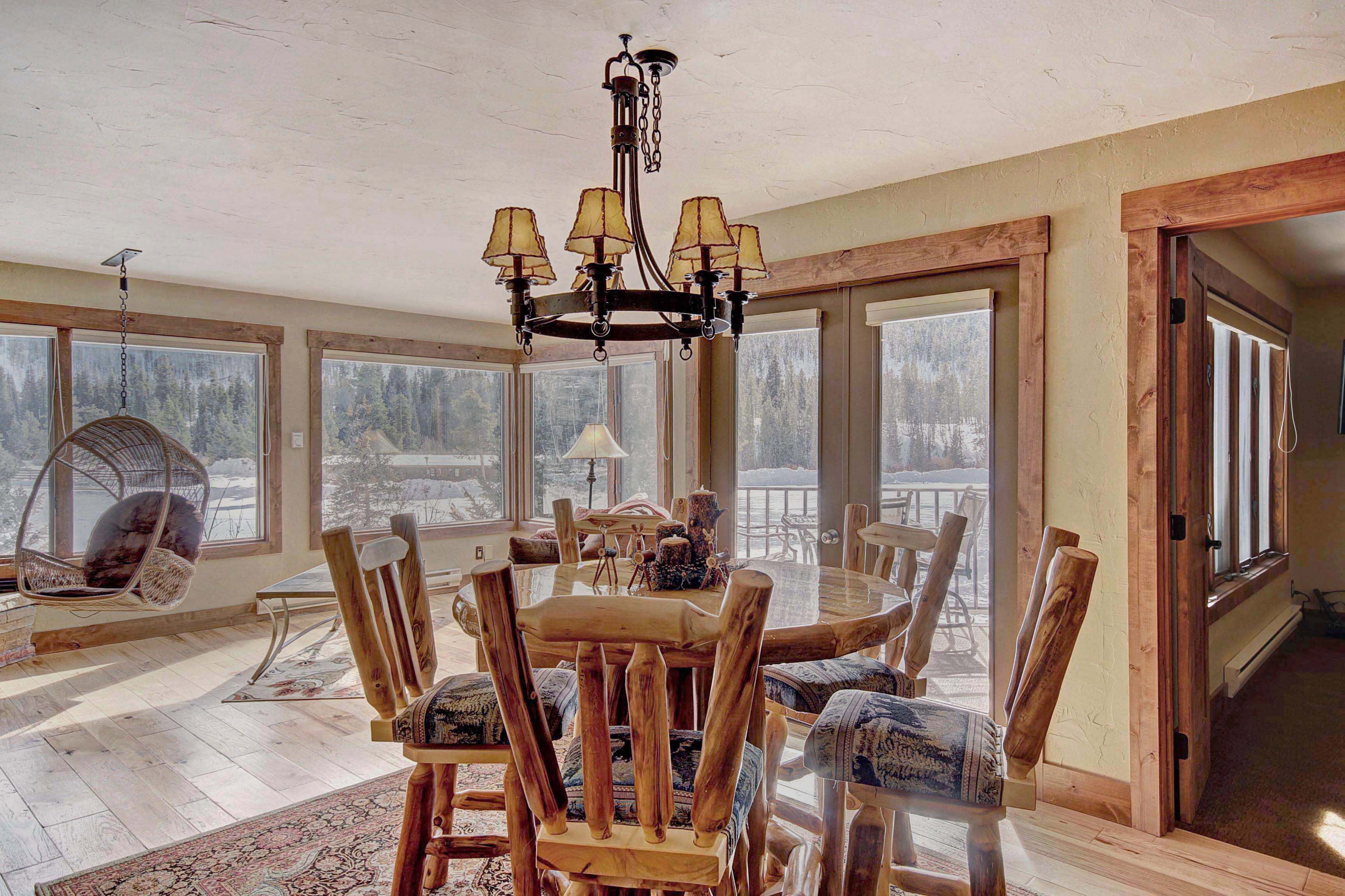 A spacious dining area with a wooden table and chairs, a chandelier, and large windows offering a view of the outdoors.