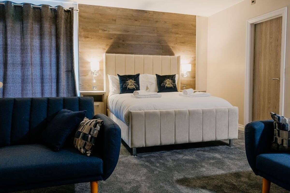 Quarto de hotel com cama, duas mesas de cabeceira, um sofá azul e uma parede de destaque em madeira.