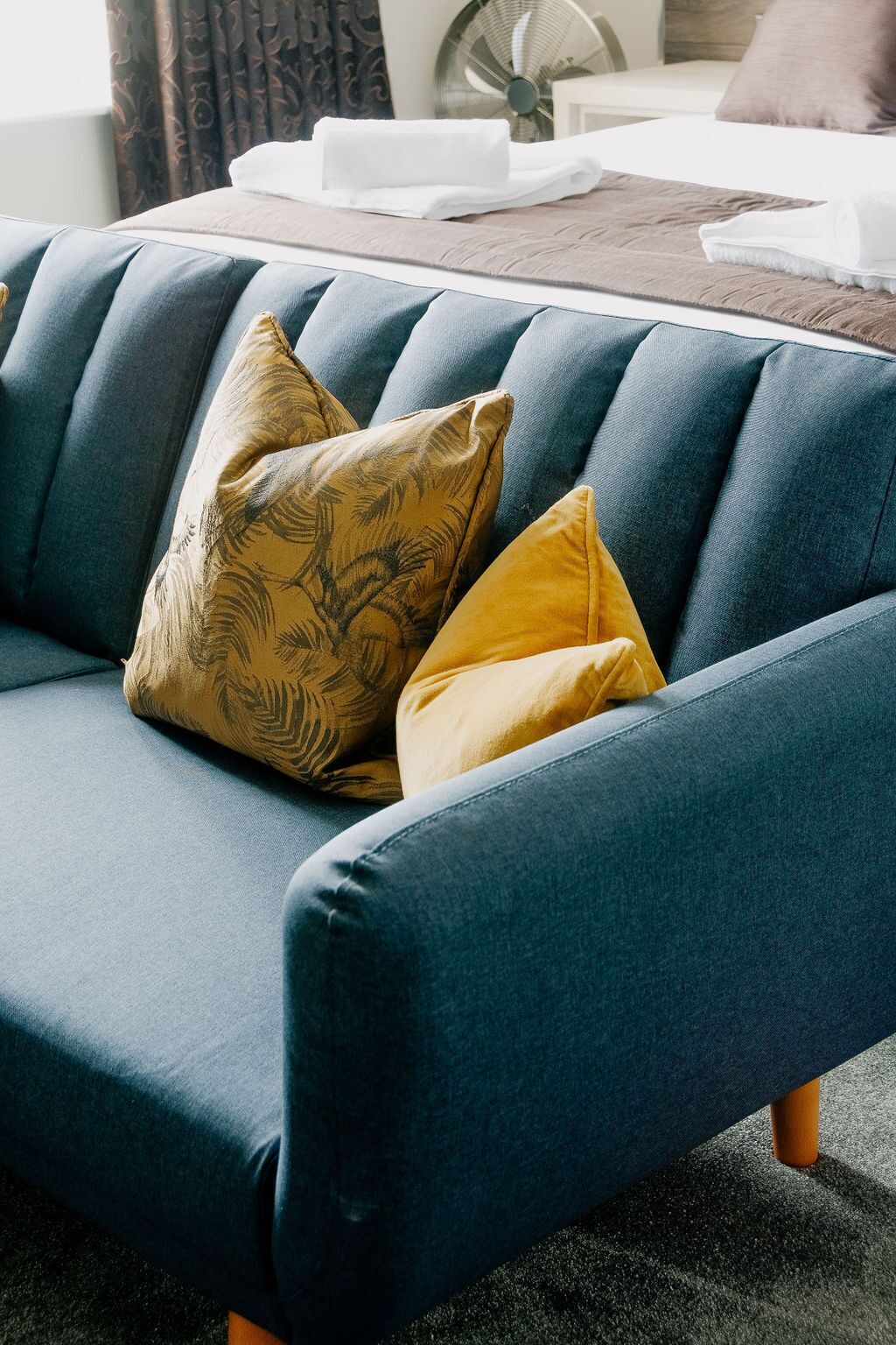 A dark blue sofa with two cushions, one with a leaf pattern and one solid yellow, in a room with a bed and a fan in the background.