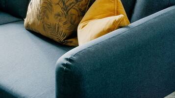 A dark blue sofa with two cushions, one with a leaf pattern and one solid yellow, in a room with a bed and a fan in the background.