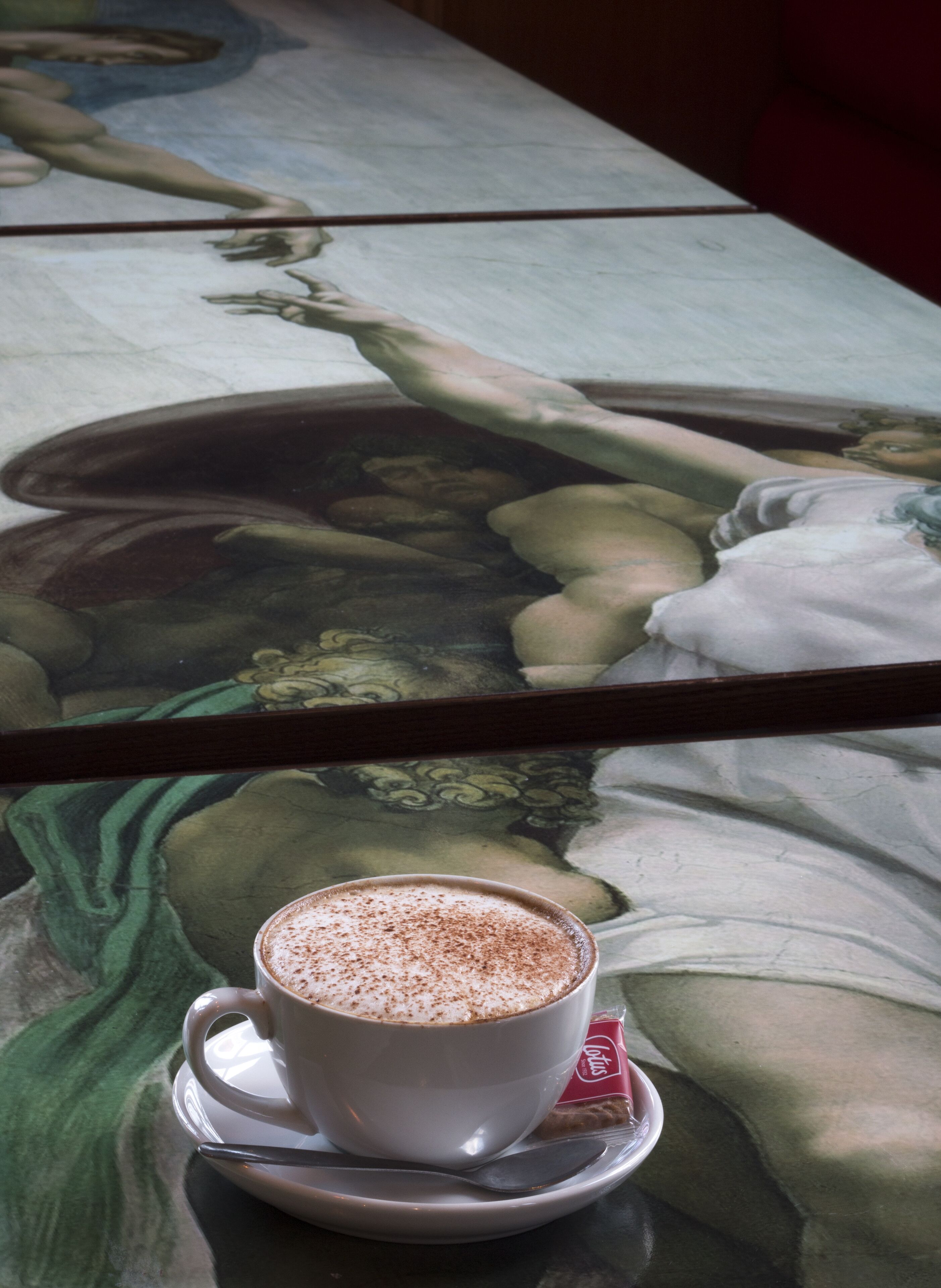 Une tasse de café posée sur une soucoupe, avec une cuillère, le tout placé sur une table devant laquelle on aperçoit un tableau.