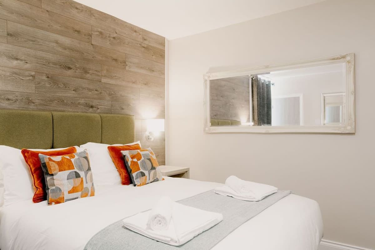 Cama bem arrumada com lençóis brancos, cabeceira verde e almofadas decorativas. Um espelho está fixado na parede acima da cama.