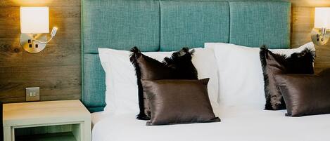 Een opgemaakt bed met een blauwgroen hoofdbord, twee bedlampen en een houten nachtkastje met een lade.