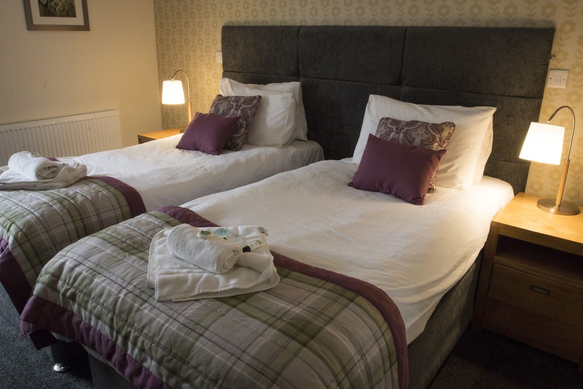 Quarto de hotel com duas camas, cada uma com cabeceira, almofadas e abajures. Há toalhas nas camas e uma mesa de cabeceira de madeira com gaveta.