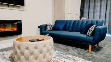 A modern living room with a blue sofa, a round ottoman, a fireplace, and a television.