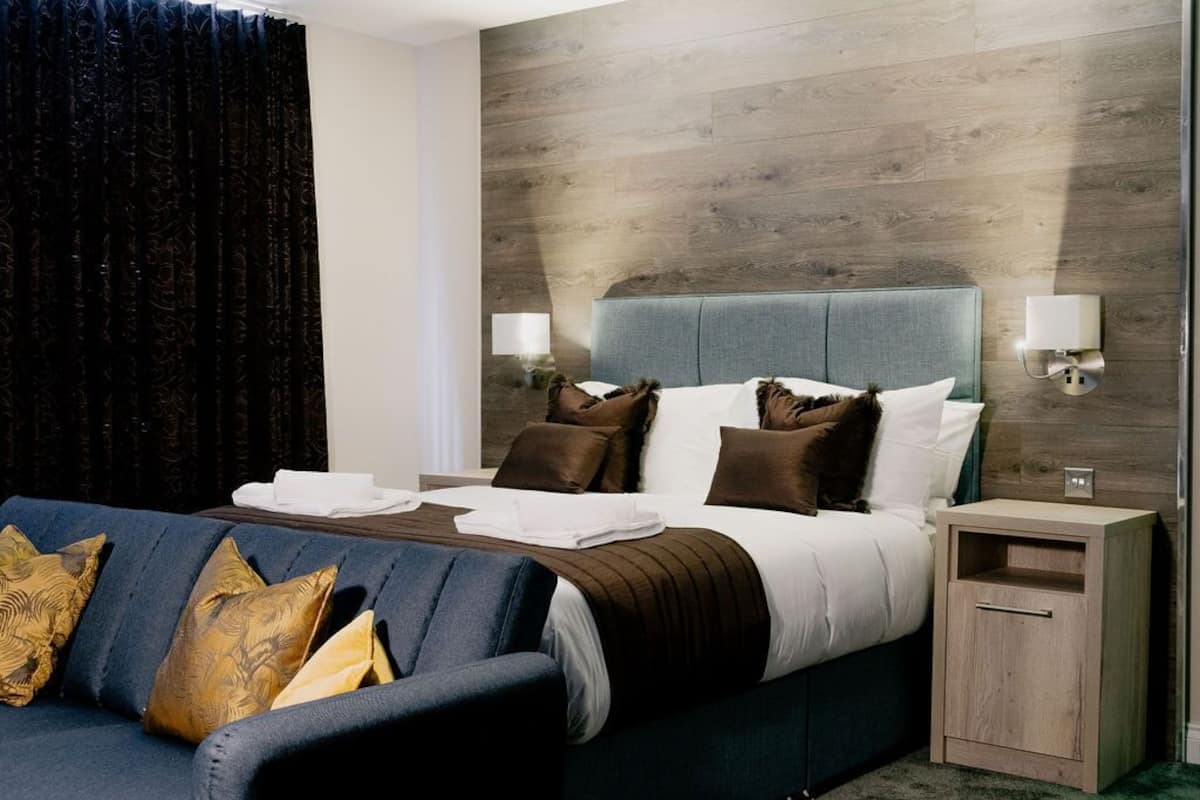 Quarto de hotel moderno com uma cama grande, um sofá azul e uma parede de destaque em madeira.