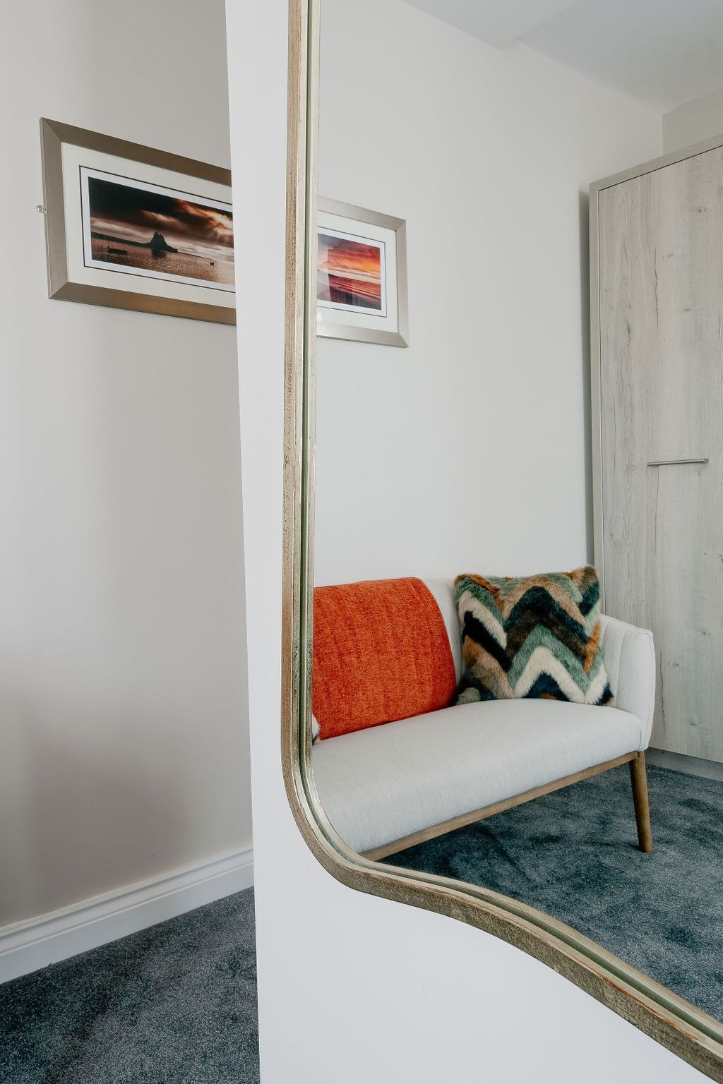 A room with a white sofa, two pillows, and a mirror reflecting a door and a framed picture on the wall.