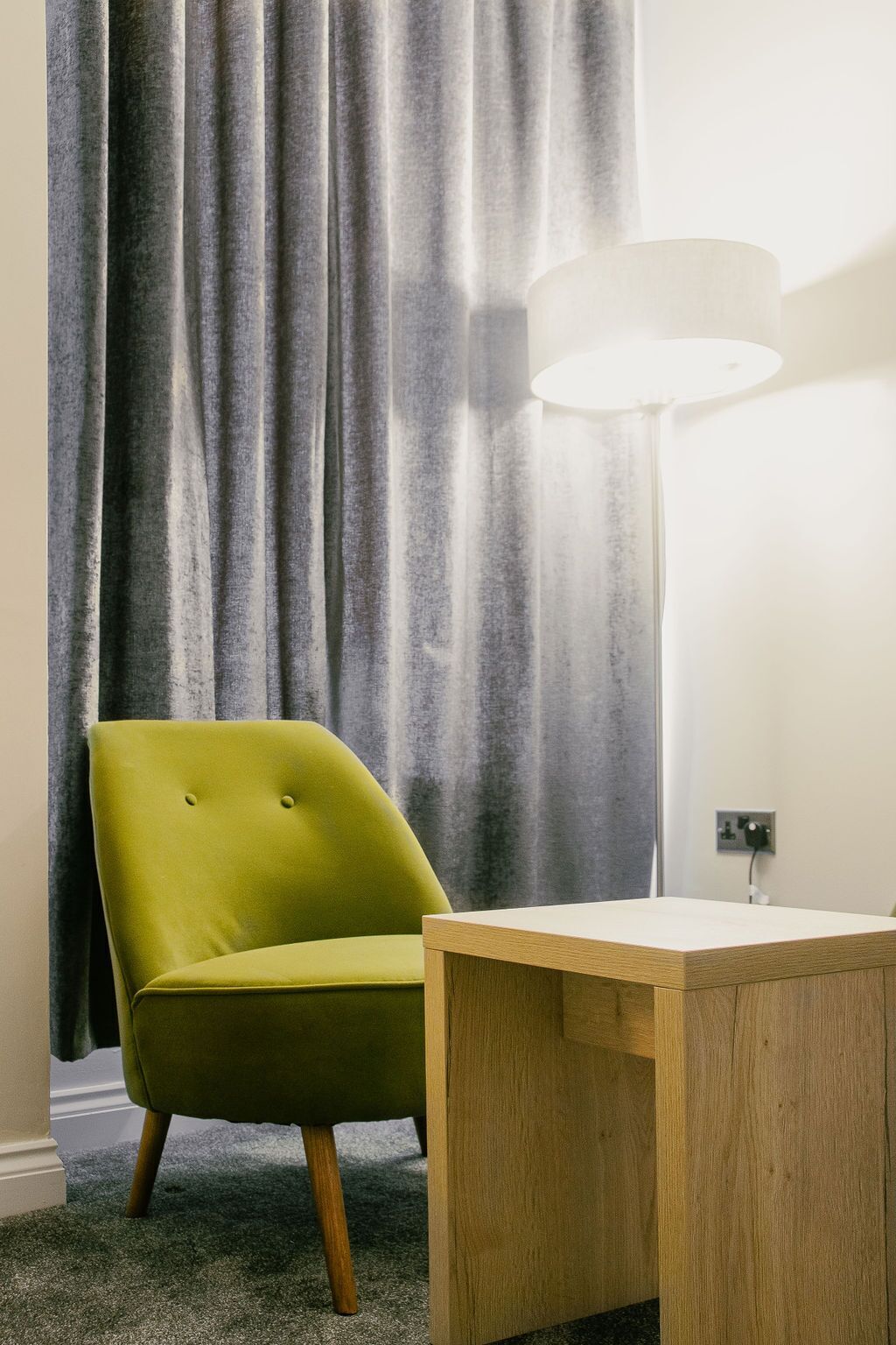A room with a green armchair, a wooden side table, a floor lamp, and a grey curtain.