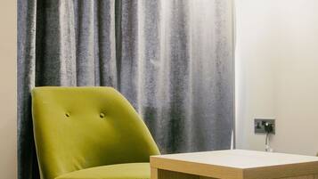 A room with a green armchair, a wooden side table, a floor lamp, and a grey curtain.