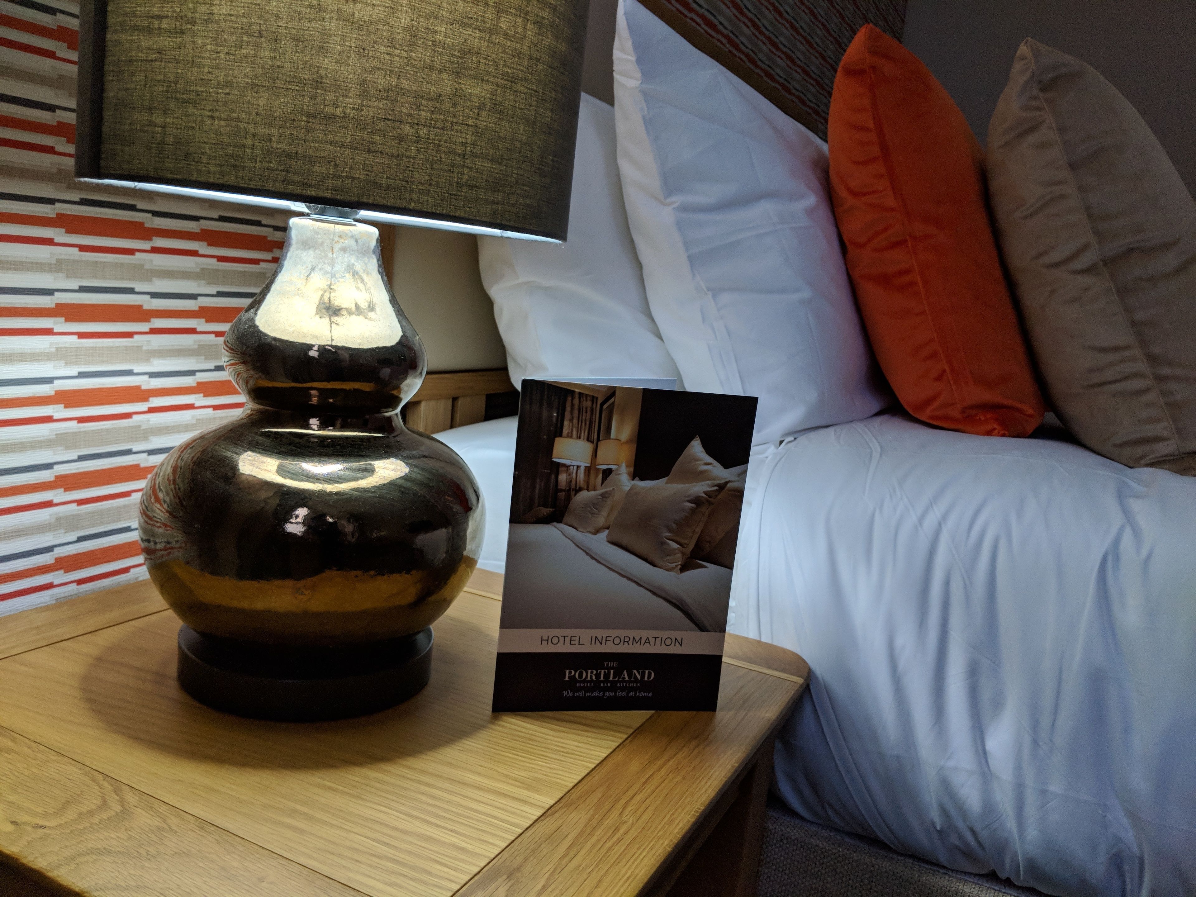 A hotel room with a wooden nightstand, a decorative vase, a framed photo of a bedroom, and a pillow with an orange accent.