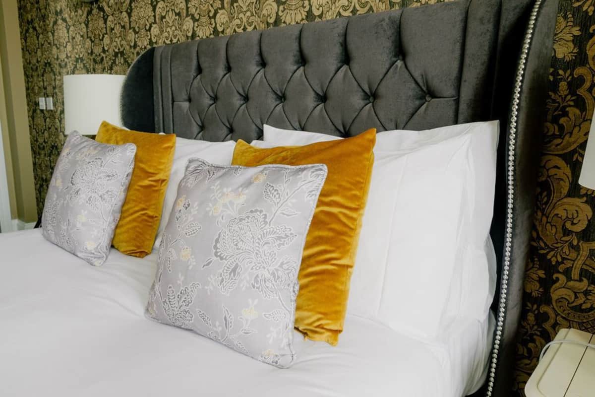 Uma cama com cabeceira estofada, várias almofadas em tons de cinza e mostarda, e uma mesinha de cabeceira branca com um abajur.