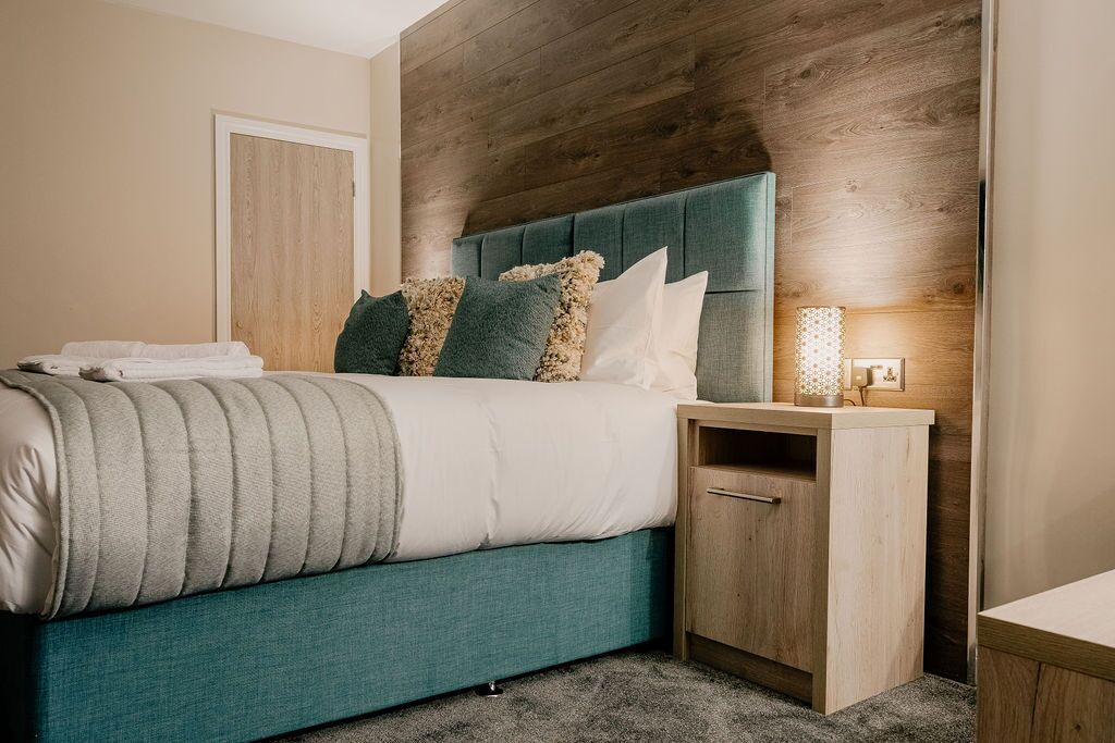 A neatly made bed with a teal headboard, a wooden nightstand with a drawer, and a wall-mounted lamp.