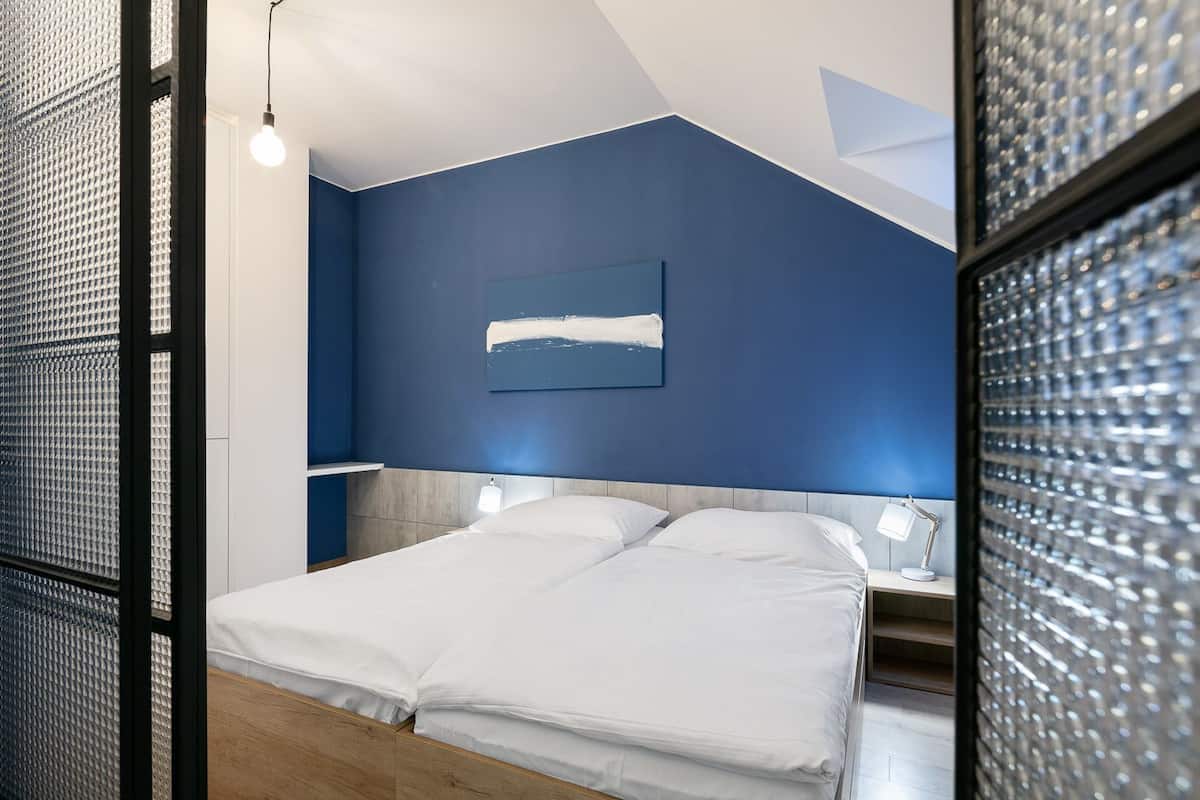 A modern bedroom with a large bed, a headboard, a bedside table with a lamp, and a wall-mounted abstract painting.