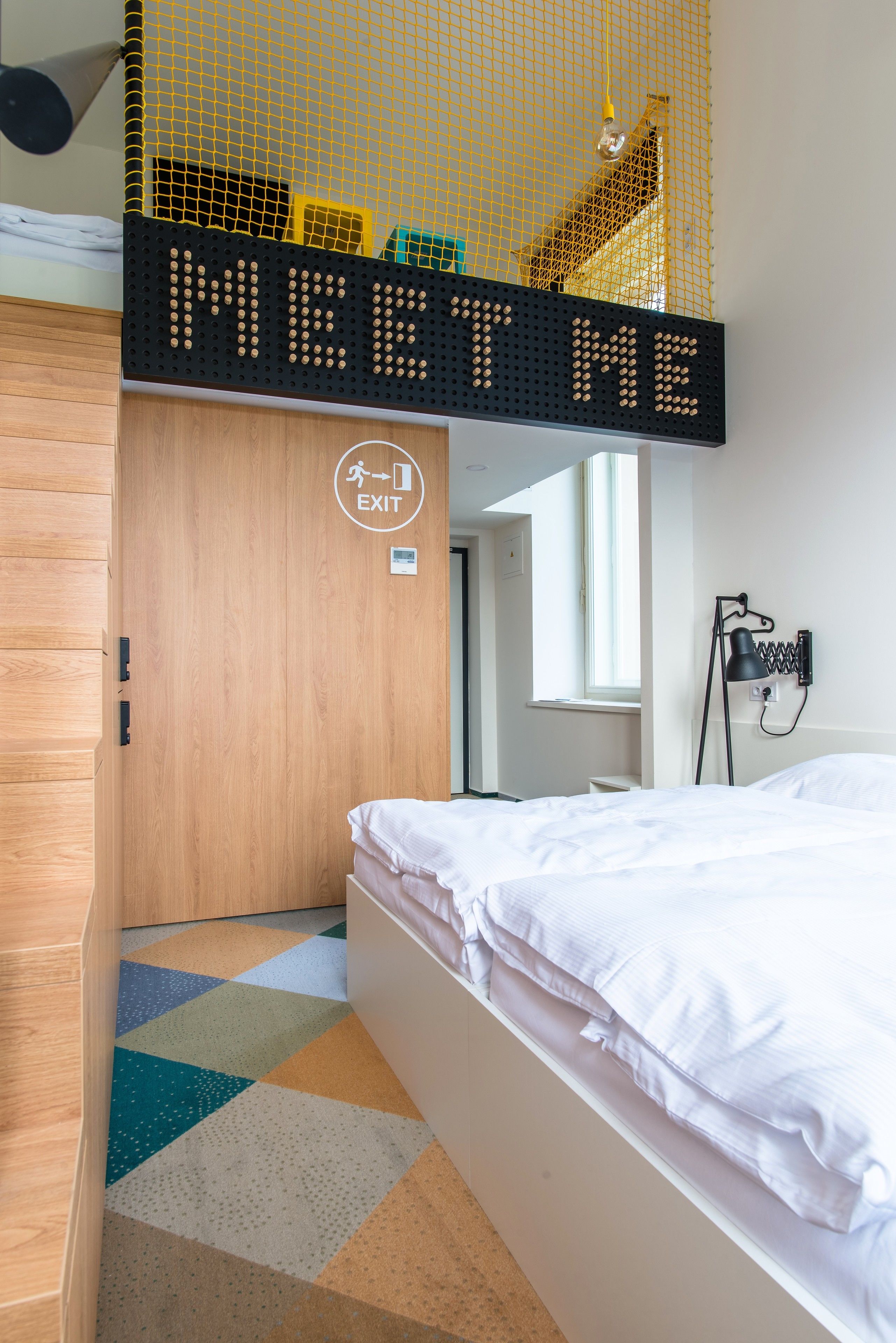 A bedroom with a bed, a sign that says "MEET ME", a door with an exit sign, and a window.