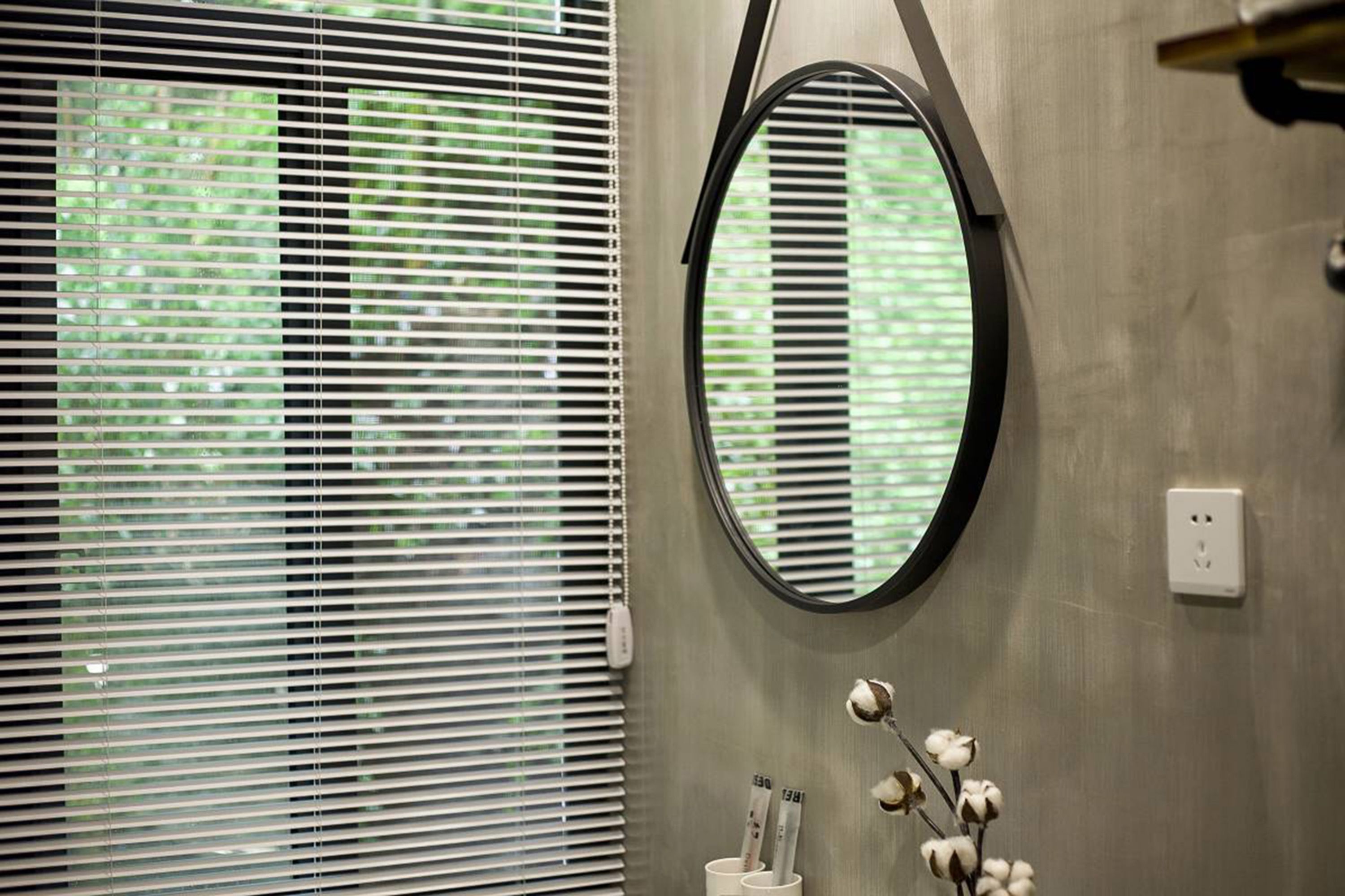 Un bagno con un'ampia finestra dotata di persiane, uno specchio rotondo e una decorazione con un ramo di cotone.