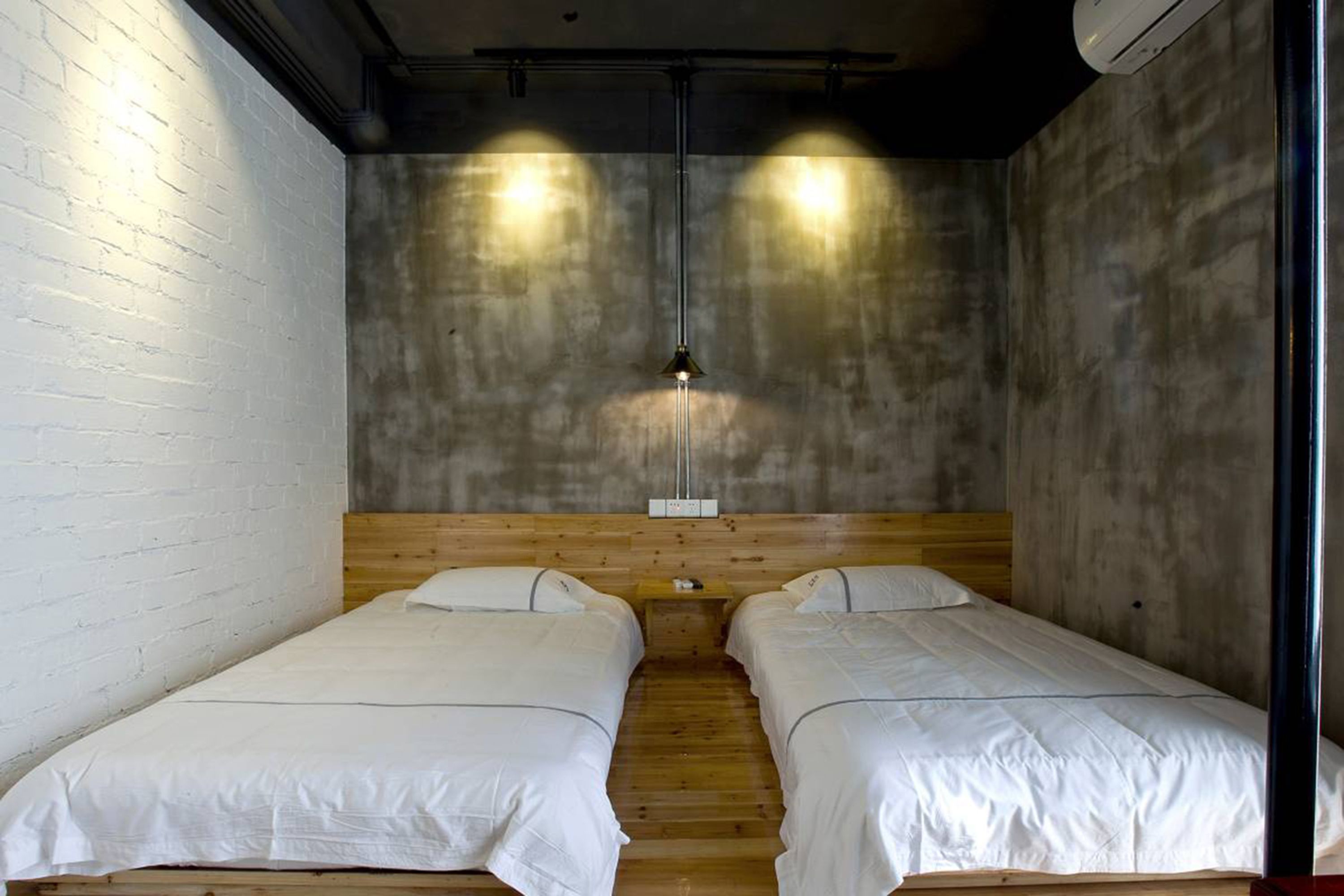 A room with two single beds, a wooden headboard, a concrete wall, and a hanging light fixture.