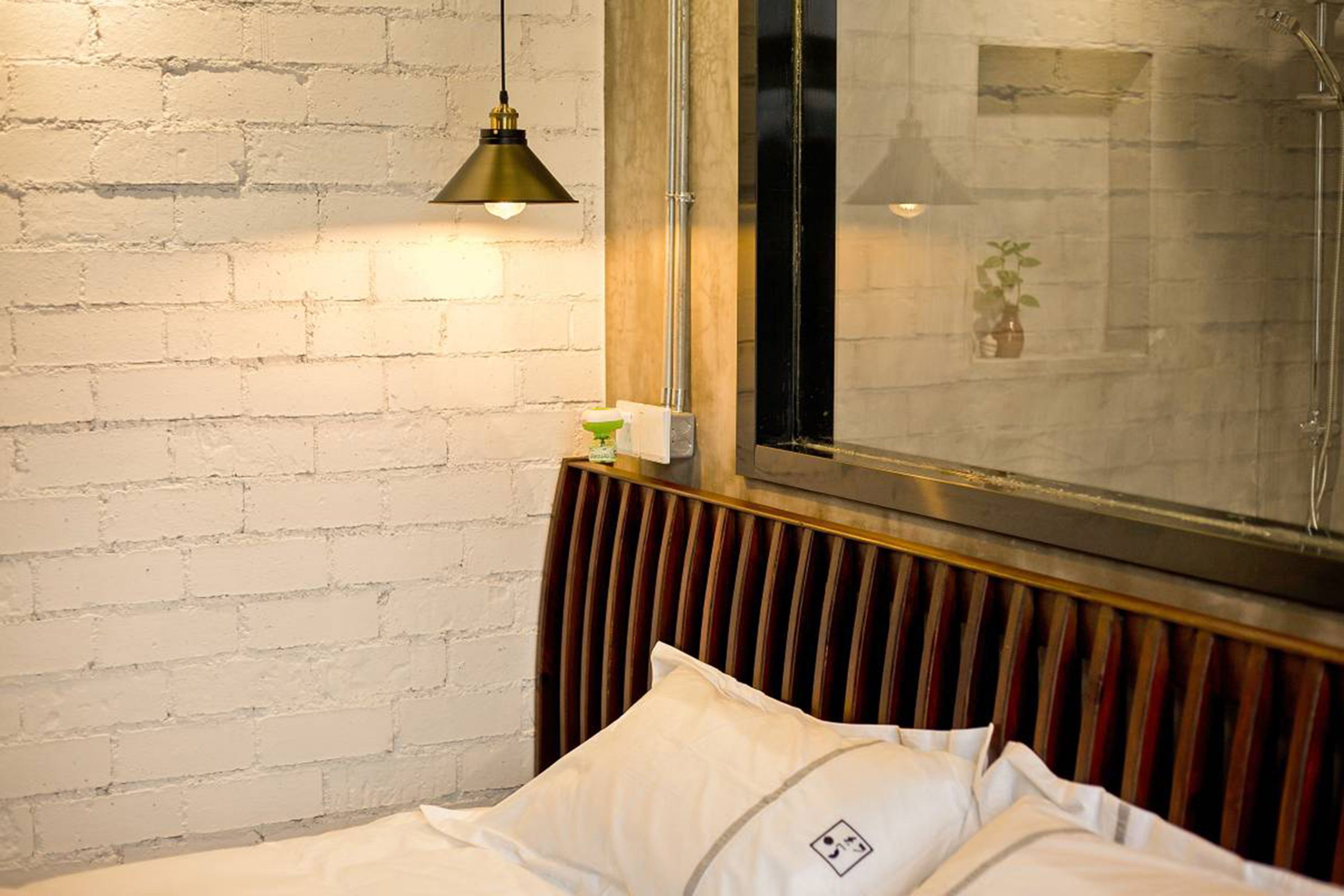 Quarto de hotel com parede de tijolos brancos, cabeceira de madeira, cama com roupa de cama branca, luminária pendente e janela com vista para uma planta.