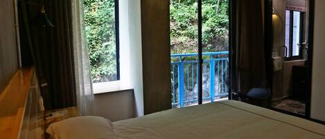 Una camera d'albergo con un letto, una finestra vista verde, un balcone con ringhiera blu e una sedia.