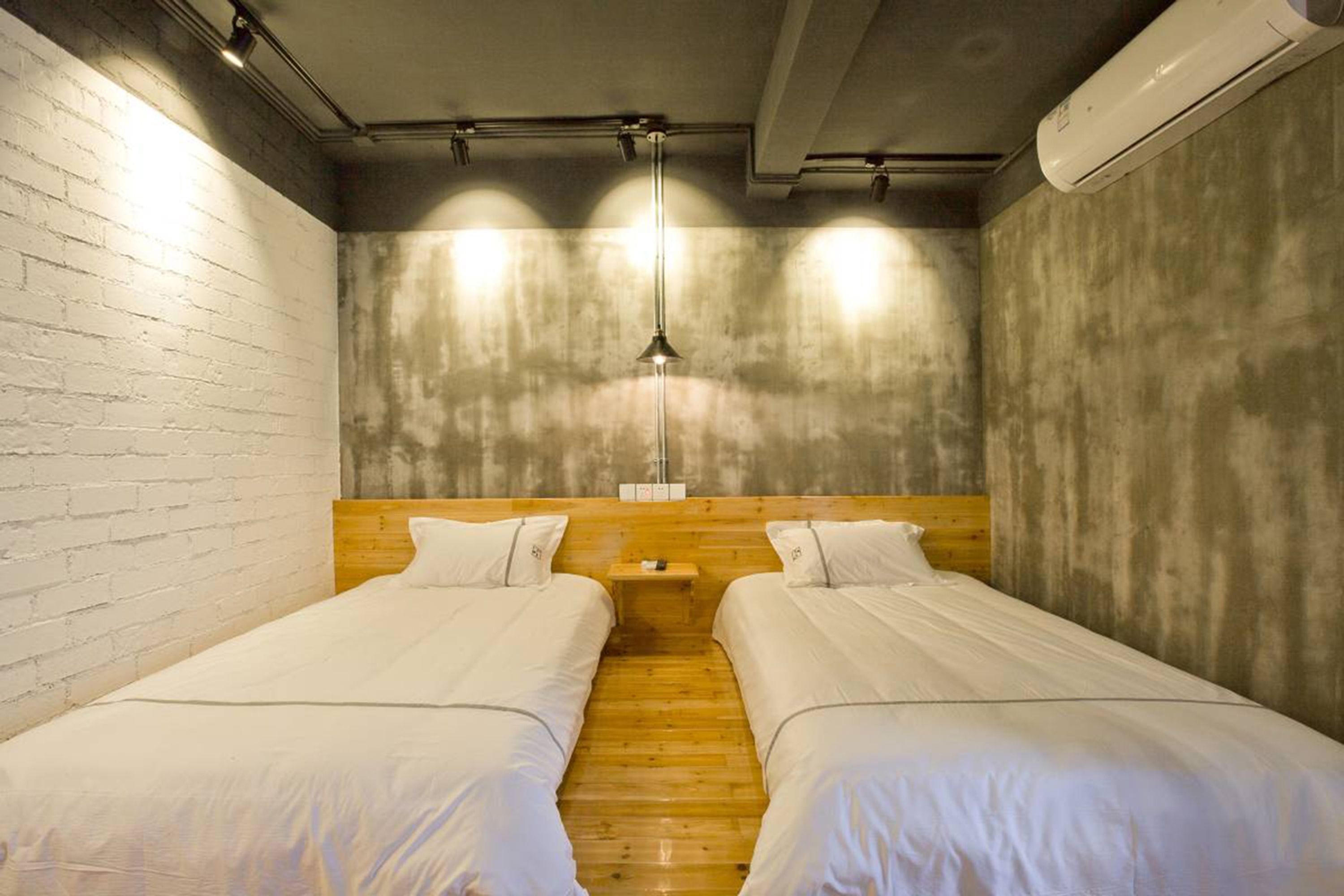A room with two single beds, wooden headboards, a small bedside table, a hanging light fixture, and an air conditioning unit.
