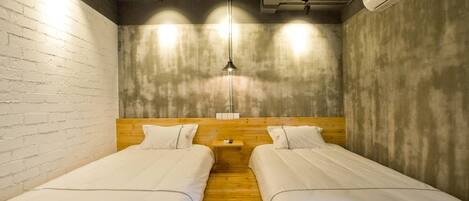 A room with two single beds, wooden headboards, a small bedside table, a hanging light fixture, and an air conditioning unit.