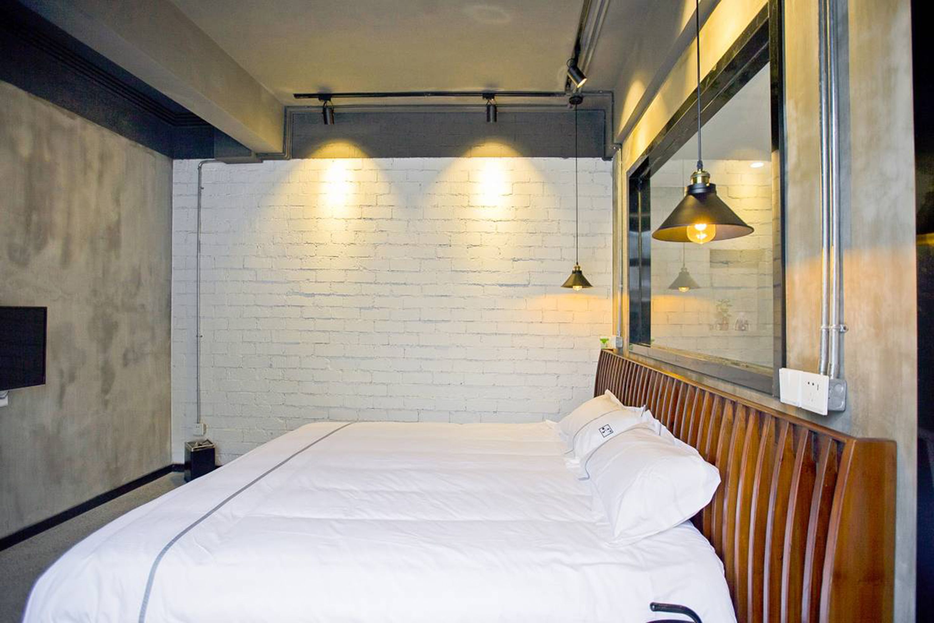A hotel room with a white brick wall, a wooden headboard, a bed with white bedding, and two pendant lights.