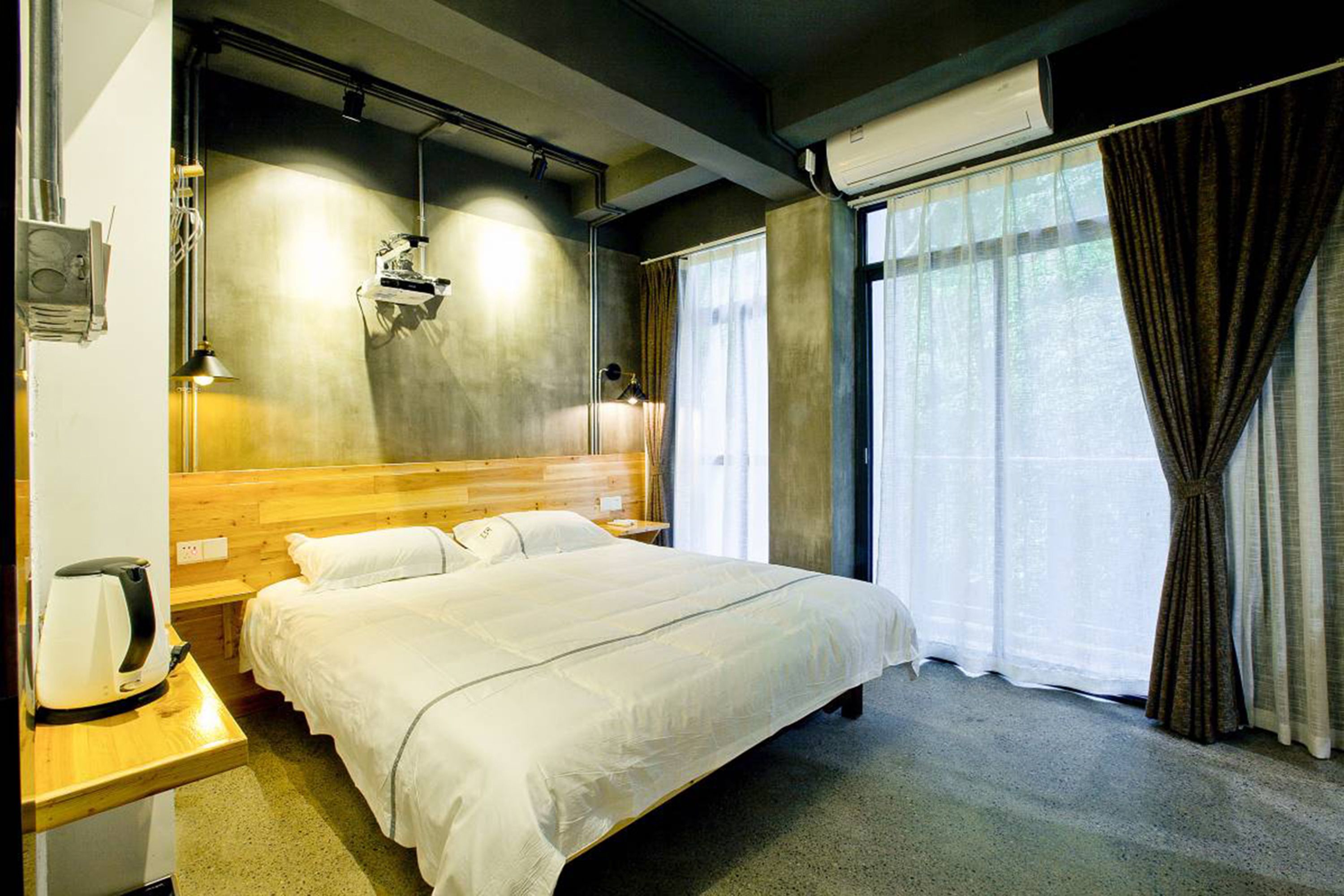 Ein Hotelzimmer mit einem hölzernen Kopfteil, einem Bett mit weißer Bettwäsche, einem Nachttisch mit Lampe und einem Fenster mit Vorhängen.