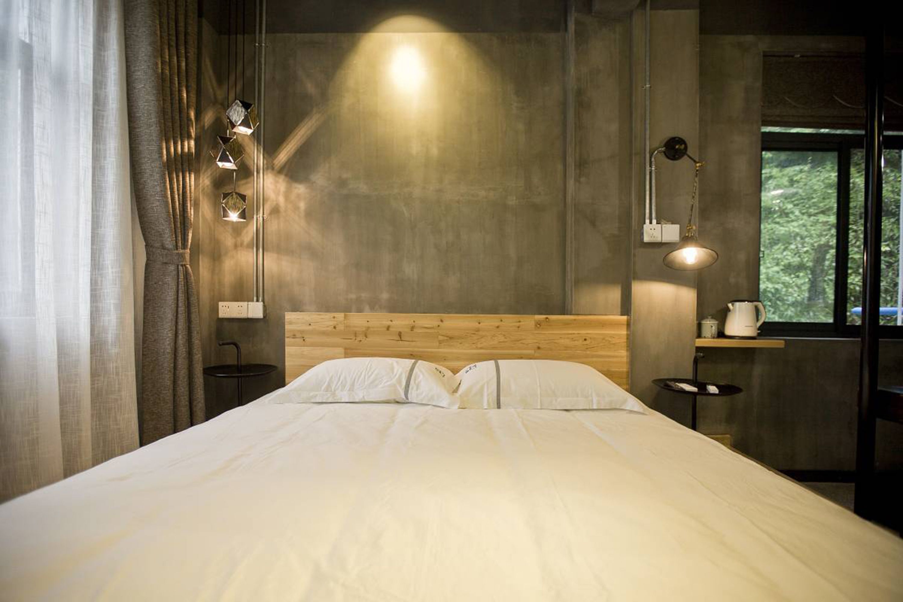 A bedroom with a wooden headboard, a bed with white linens, a bedside table with a lamp, and a window with a view of greenery.