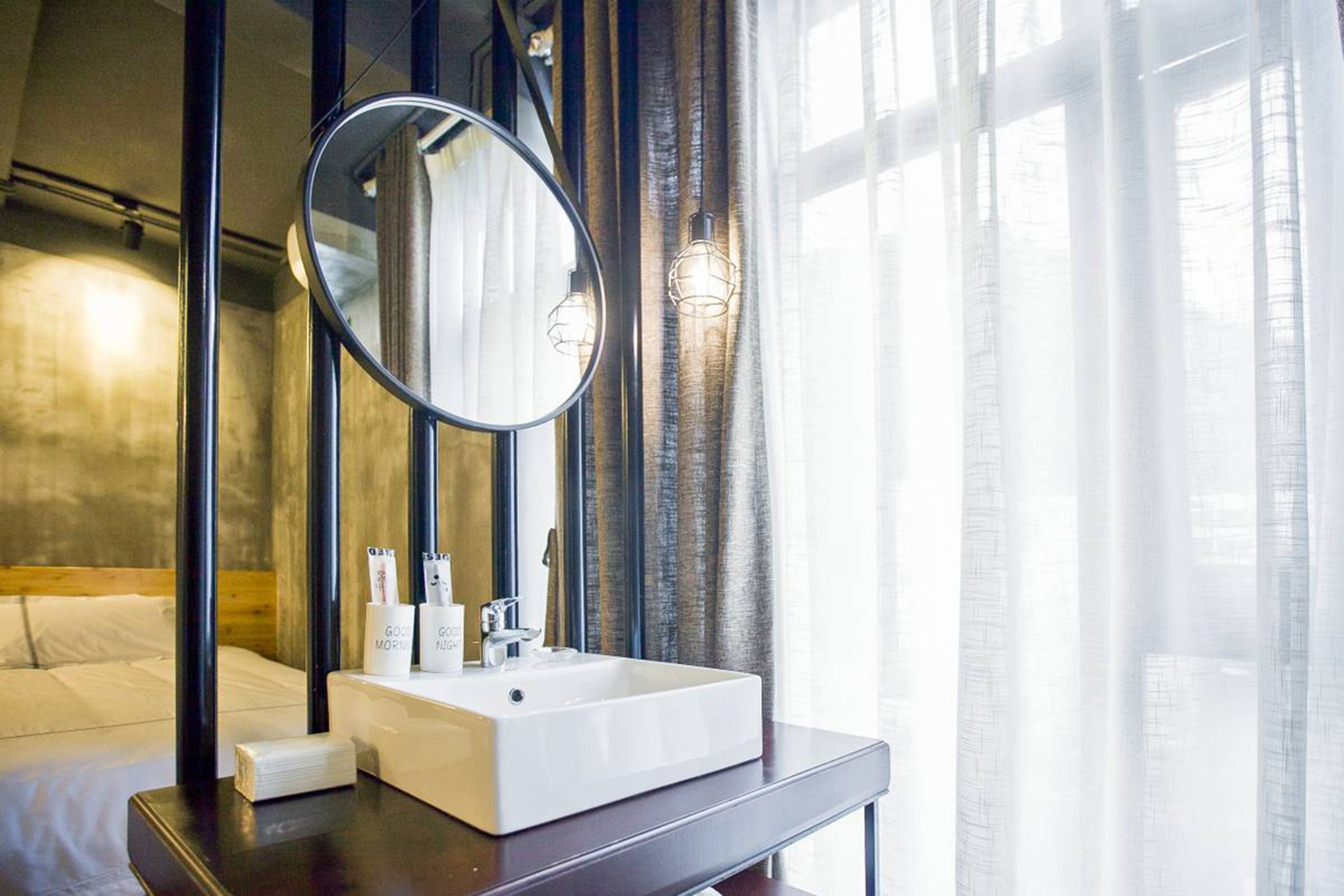A modern bathroom with a large mirror, white sink, and a bedside table with toiletries.
