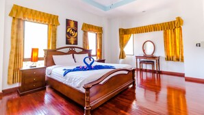 Une chambre spacieuse avec un lit en bois, de grandes fenêtres avec des rideaux, un ventilateur de plafond et une table de chevet avec une lampe.