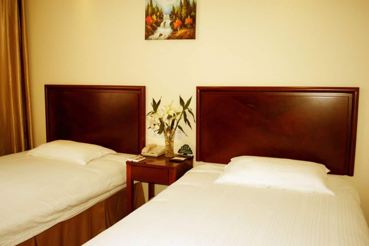 Habitación de hotel con dos camas individuales, cabecera de madera, mesita de noche con un jarrón de flores y un cuadro de un paisaje montañoso.