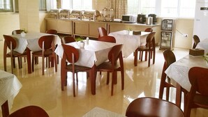A dining area with tables and chairs, a kitchen in the background, and large windows with curtains. - GreenTree Inn Taizhou Xinghua Anfeng Runan Garden Express Hotel (Taizhou)