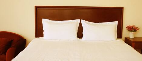 A neatly made bed with a wooden headboard, a small bedside table with a vase of flowers, and a red chair to the left.