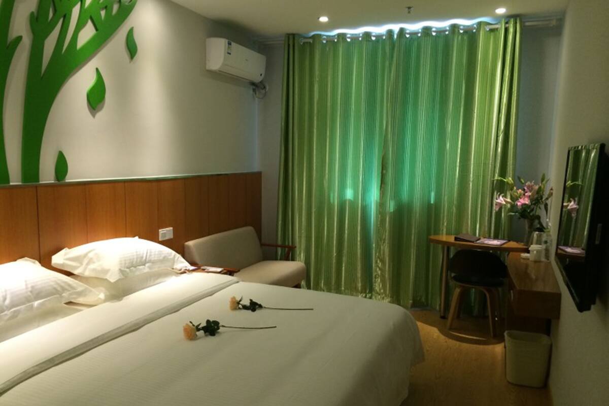 A hotel room with a bed, a desk, a chair, a trash bin, and a wall-mounted air conditioner.