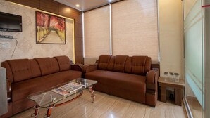 A modern living room with a brown sectional sofa, a glass coffee table, a flat-screen TV mounted on the wall, and large windows with blinds. - Eclat Suites Mint Gomti Nagar (Lucknow)
