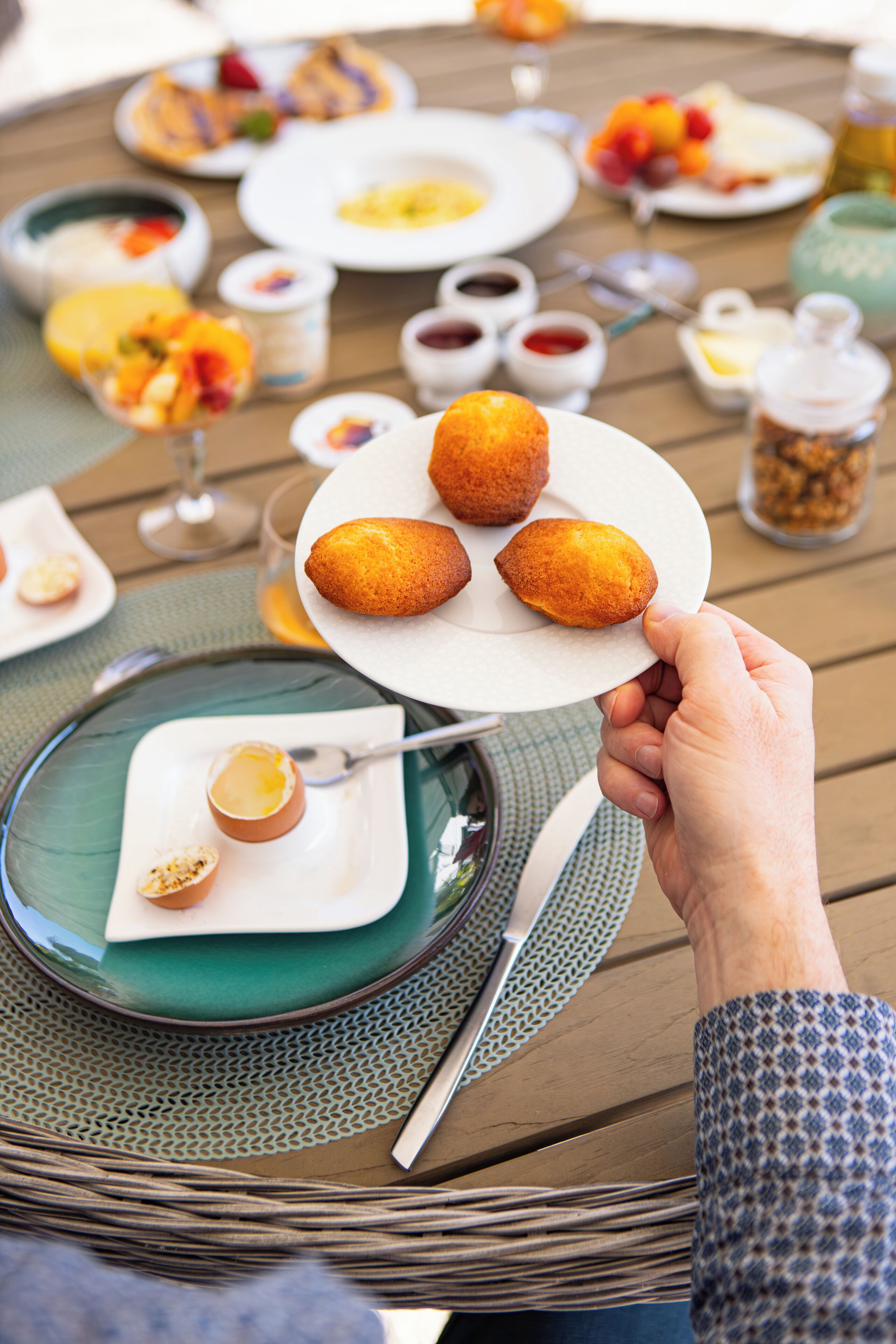 Una mesa de comedor con platos de comida, una mano sosteniendo un plato con croissants y un vaso de jugo de naranja.