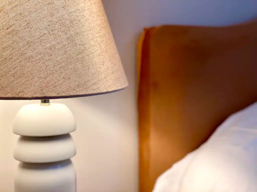 A close-up of a lamp with a textured lampshade and a white base, positioned next to a wooden headboard and white bedding.