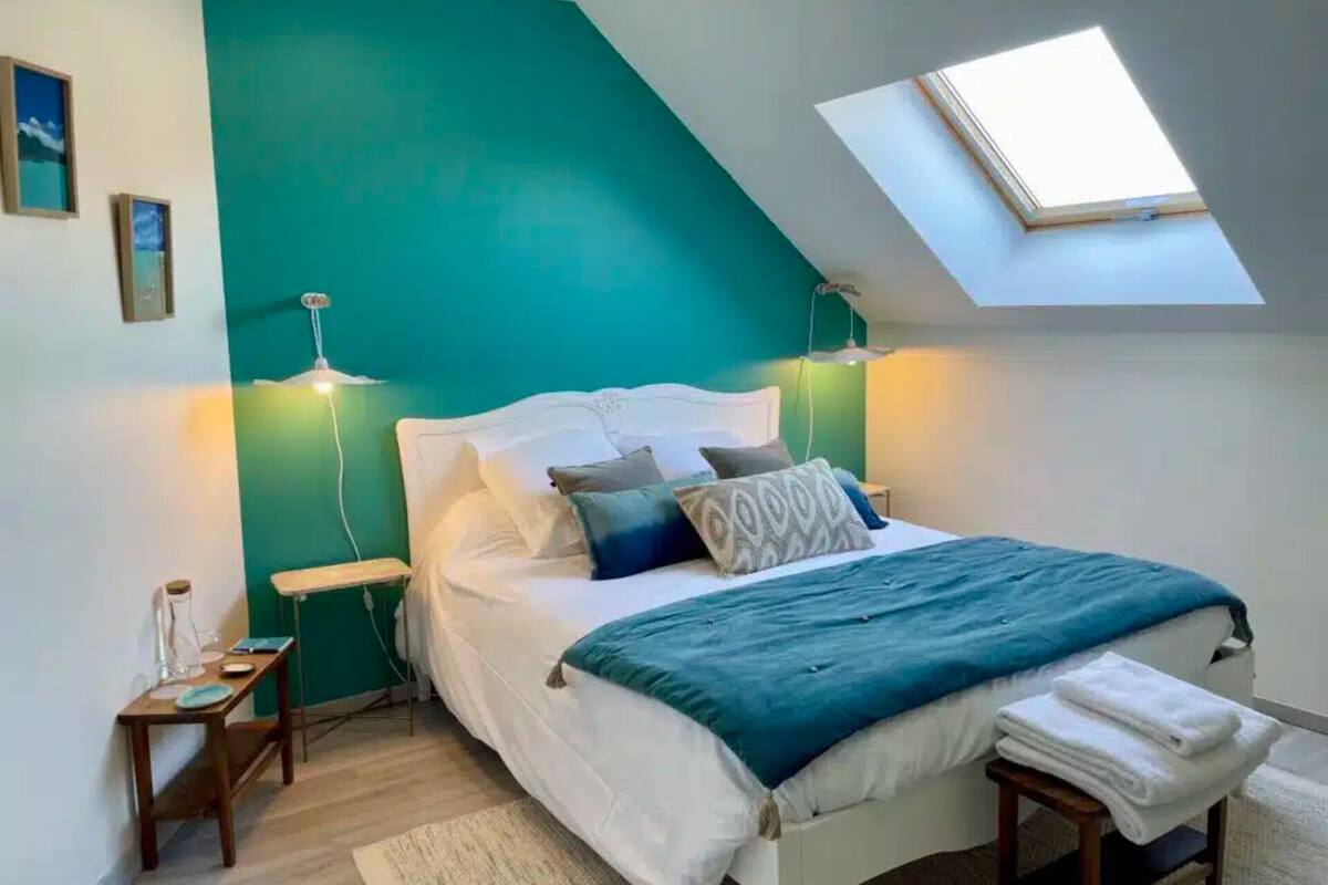 Ein Schlafzimmer mit schrägen Wänden, einem Oberlicht, einem Bett mit weißer Bettwäsche und zwei Nachttischen mit Lampen.