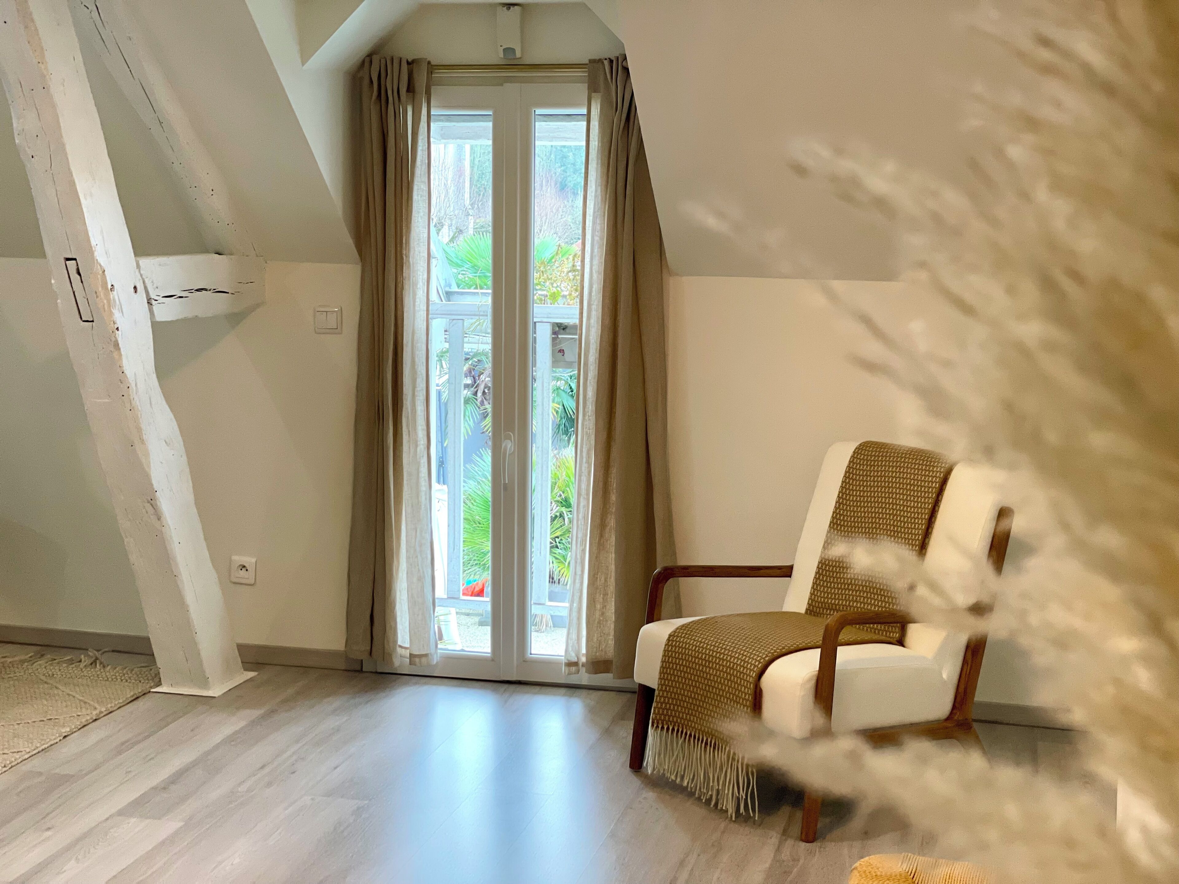 Una habitación con una silla blanca, piso de madera, un gran ventanal con cortinas y vistas a la vegetación.