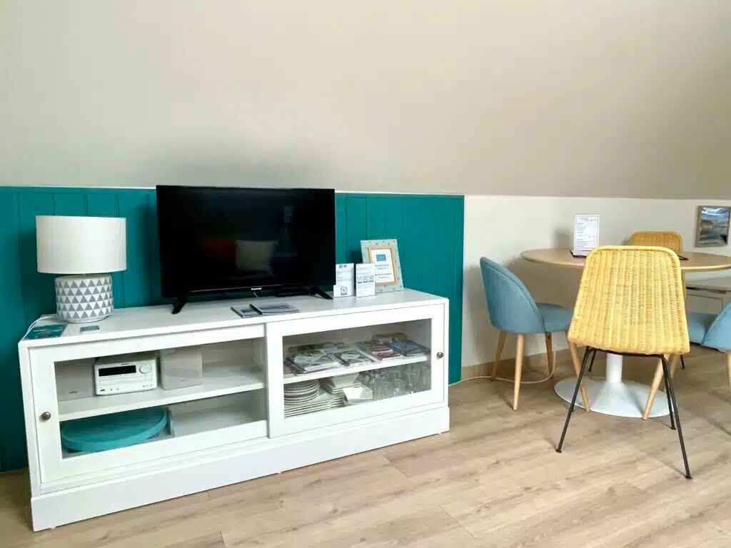 A room with a white entertainment unit, a television, a lamp, and a dining area with chairs and a table.