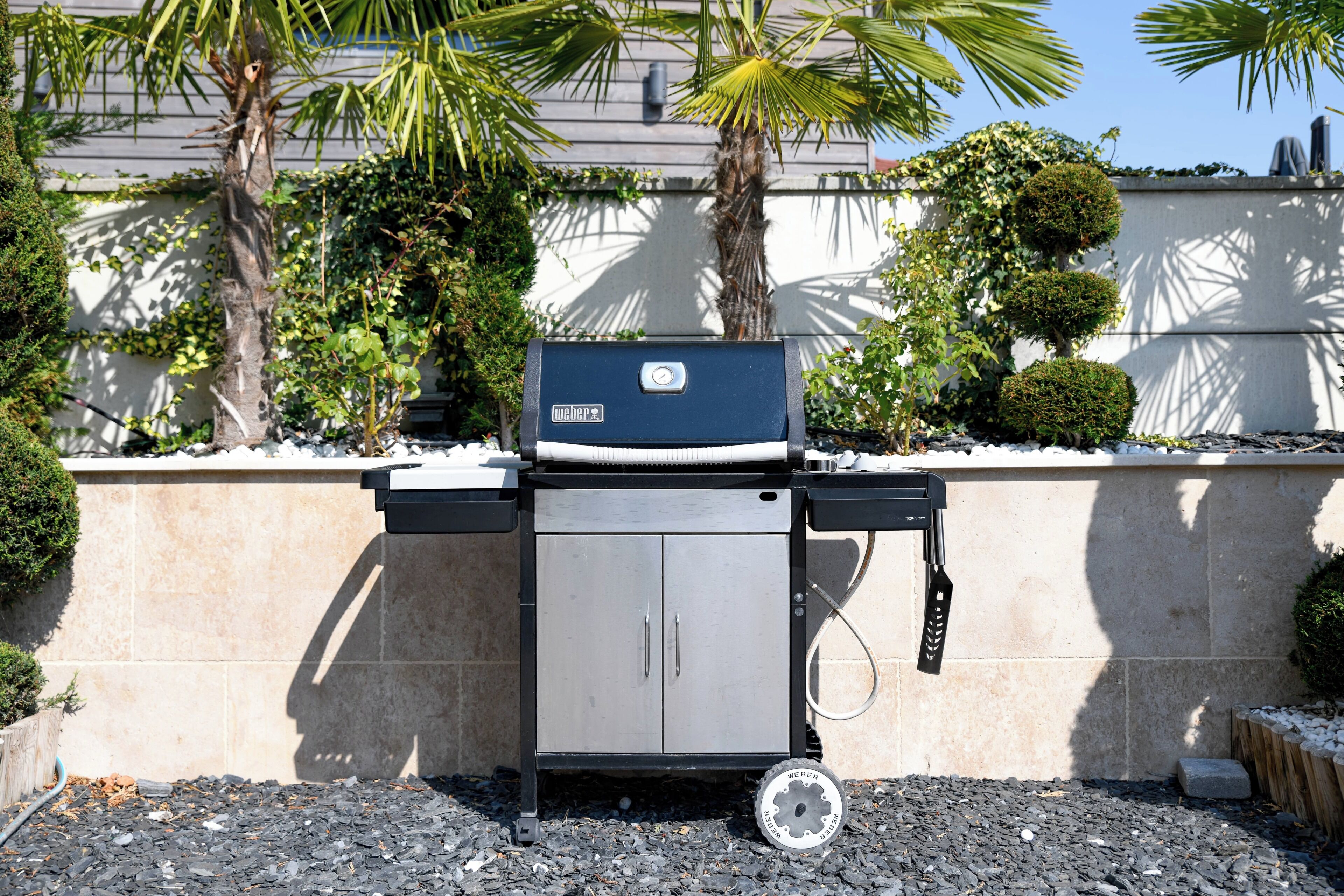 Un barbecue Weber portable, doté d’un meuble latéral et d’un tuyau, placé devant un mur de pierres et une végétation luxuriante.