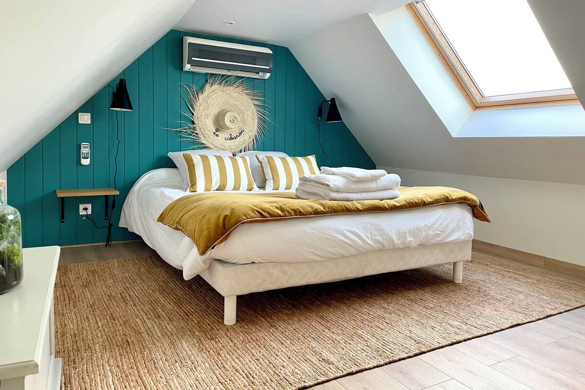 Ein Schlafzimmer mit schrägem Dach, einem Bett mit weißer Bettwäsche, einer Wand in Türkis, einem großen Fenster und einem Deckenventilator.