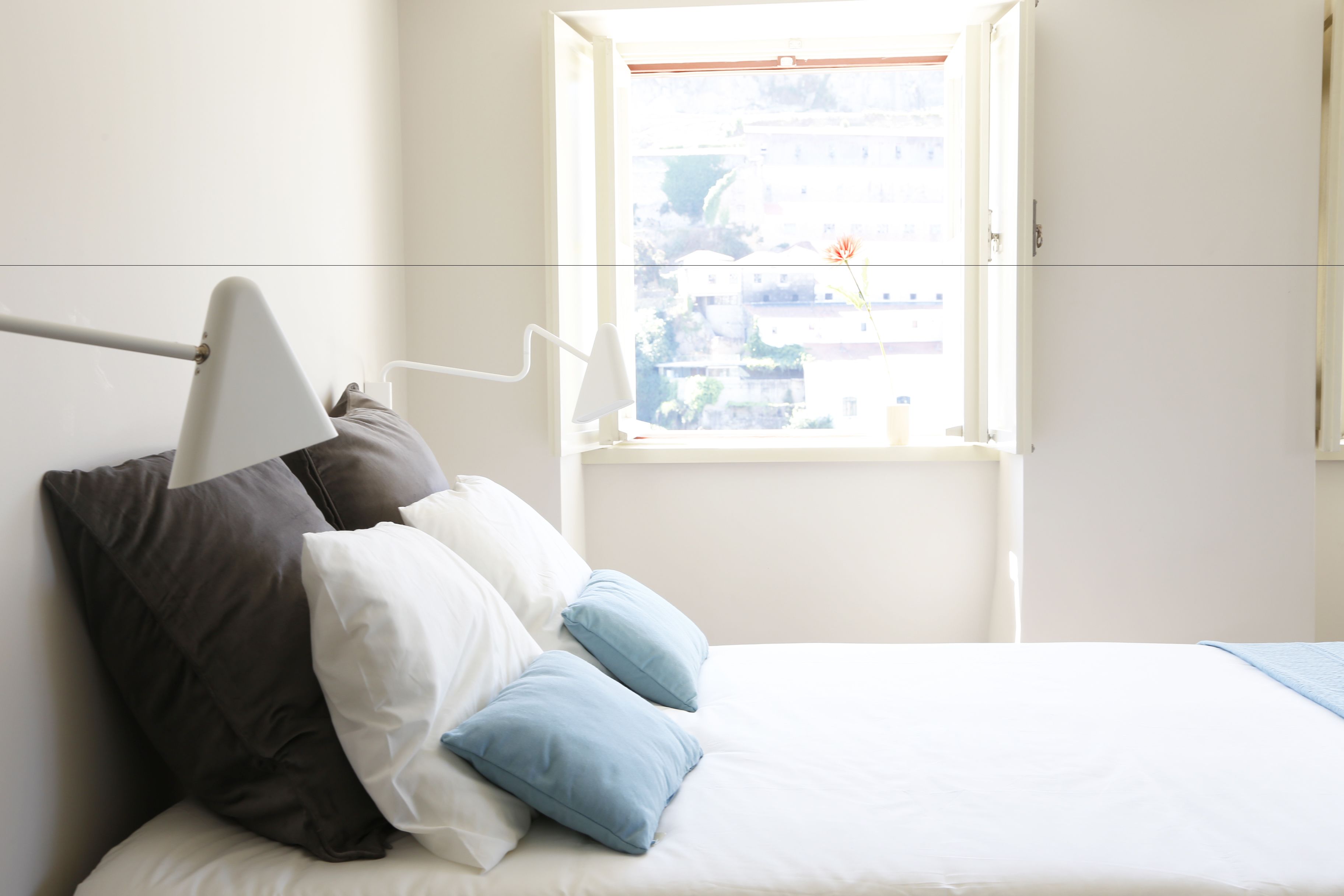 A bed with white and blue pillows, a white lamp, and a window with a view of buildings.