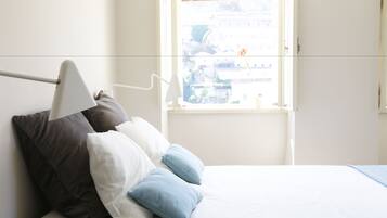A bed with white and blue pillows, a white lamp, and a window with a view of buildings.