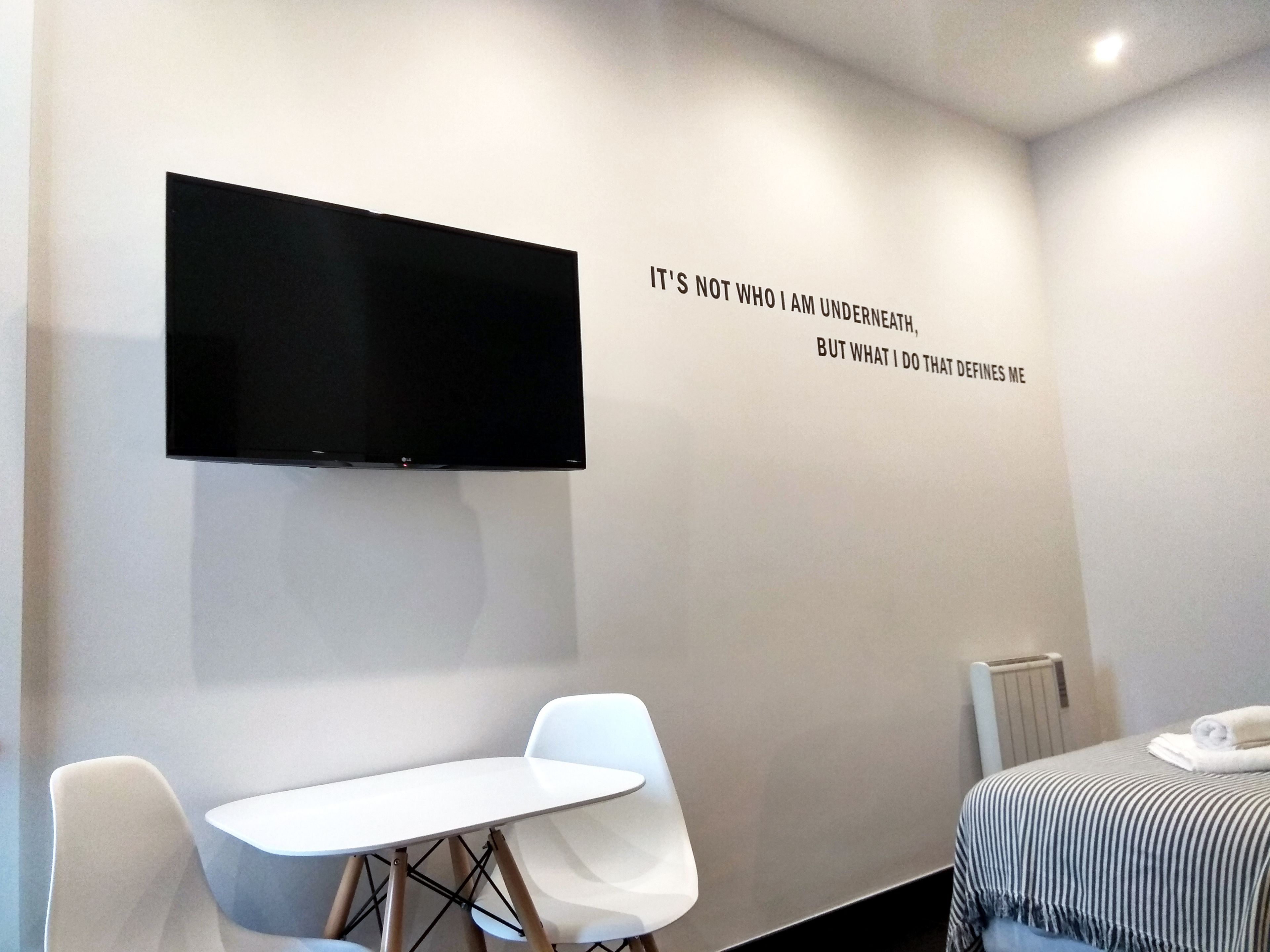 Una habitación de hotel moderna con una televisión de pantalla plana montada en la pared, una pequeña mesa blanca, una silla blanca individual y una cama con una manta a rayas.