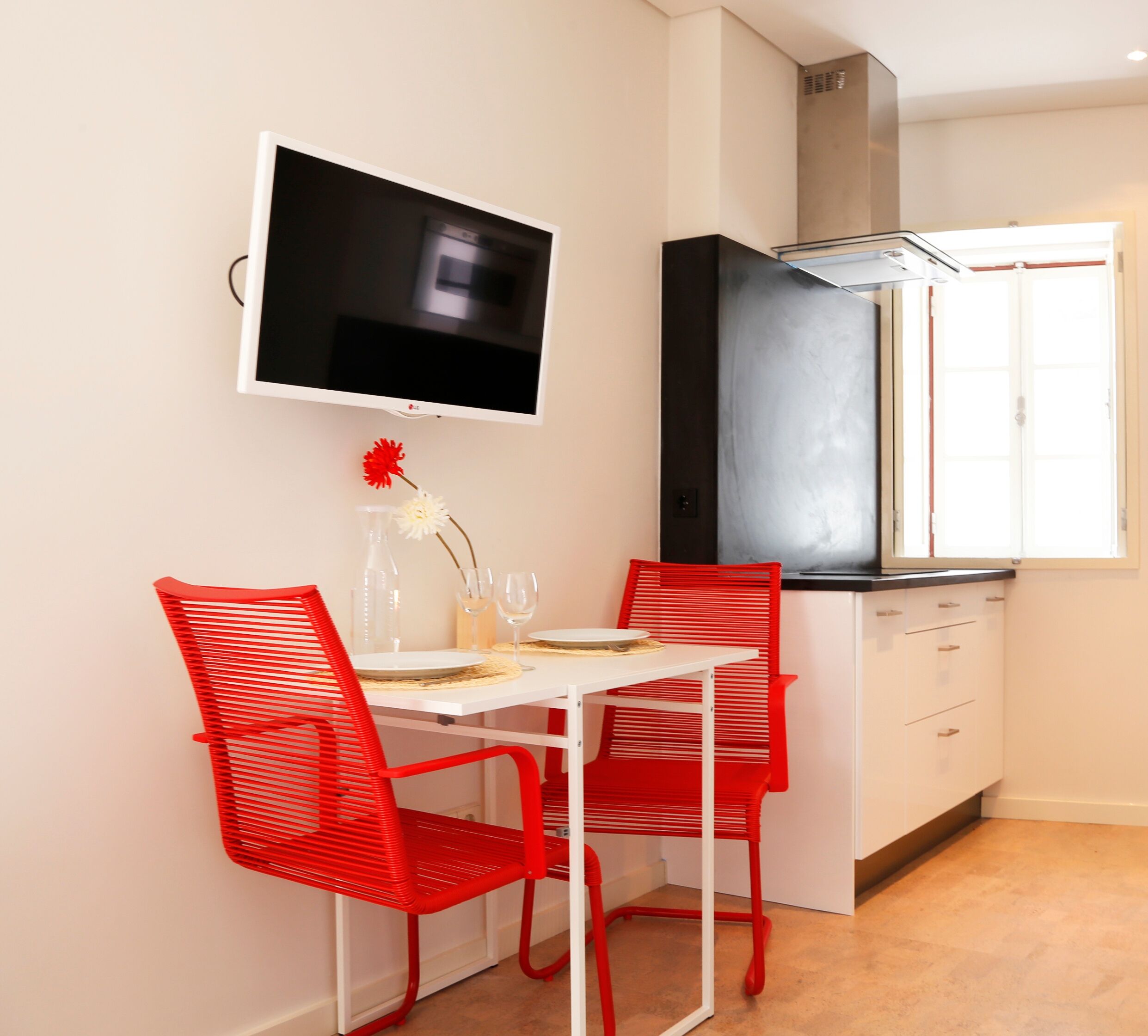 A small kitchen with a white table and red chairs, a flat-screen TV mounted above the table, and a window with a view of the outside.