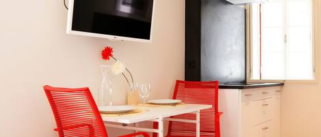 A small kitchen with a white table and red chairs, a flat-screen TV mounted above the table, and a window with a view of the outside.