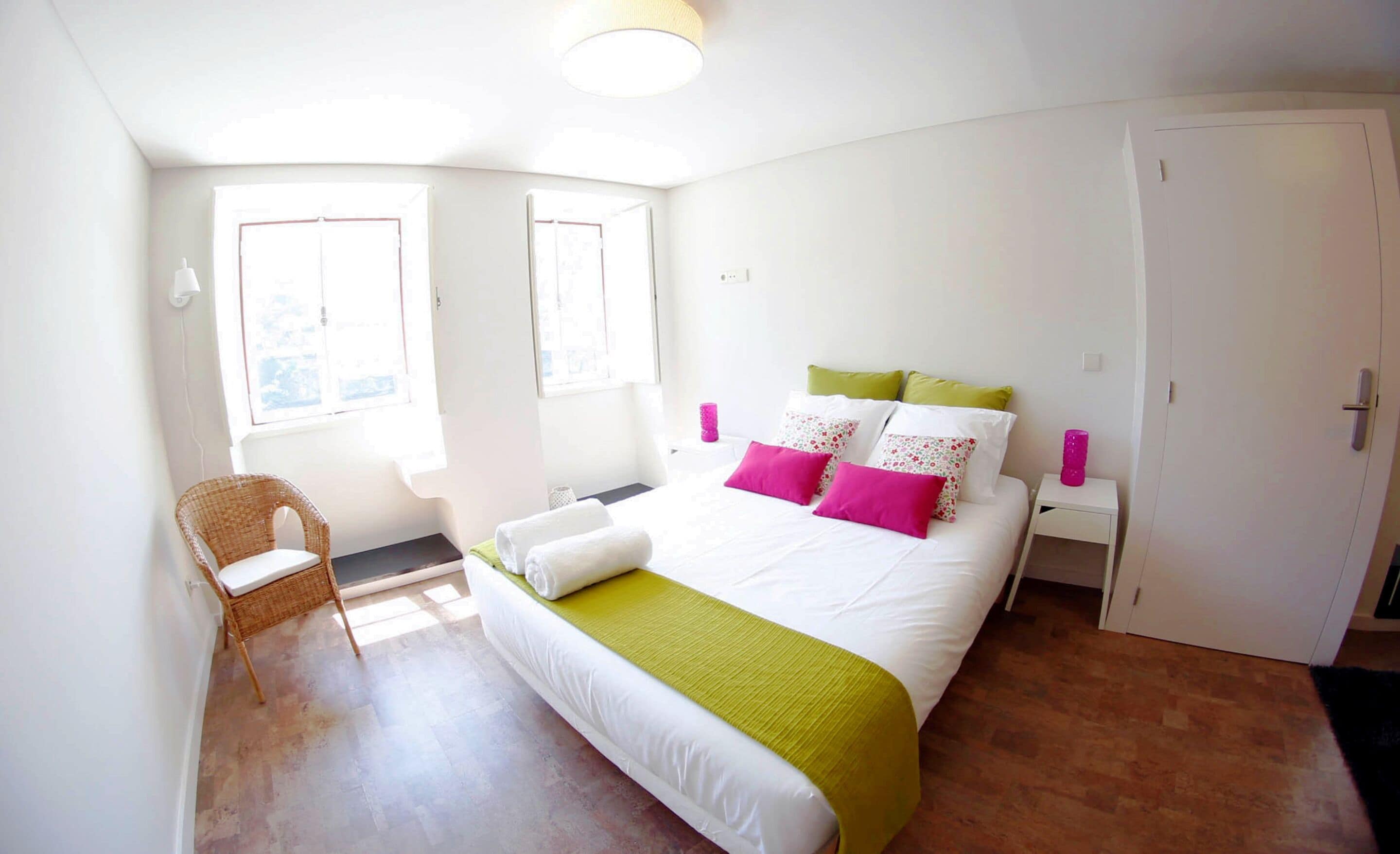 Un dormitorio con cama, una silla, armario y dos ventanas.