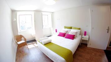 Un dormitorio con cama, una silla, armario y dos ventanas.