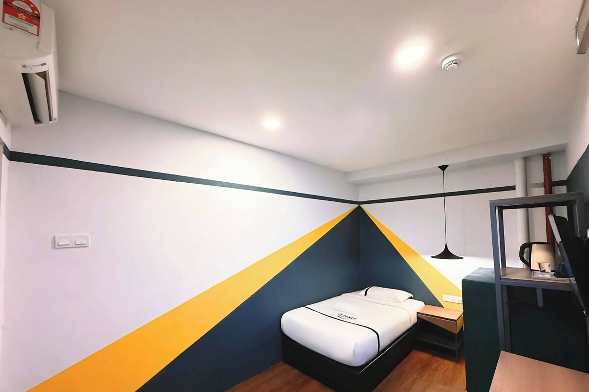 Una habitación pequeña y moderna con una cama, un escritorio y una silla. Cuenta con un aire acondicionado de pared y una lámpara en el techo.