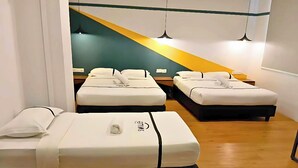 Une chambre d’hôtel avec trois lits parfaitement faits, chacun agrémenté de draps blancs et d’une tête de lit noire. On y trouve des tables de chevet avec des lampes, et le sol est en bois.